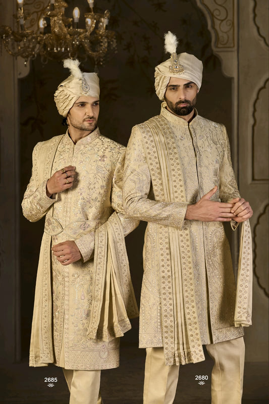 Two men in traditional cream-colored sherwanis with turbans standing against a dark background.