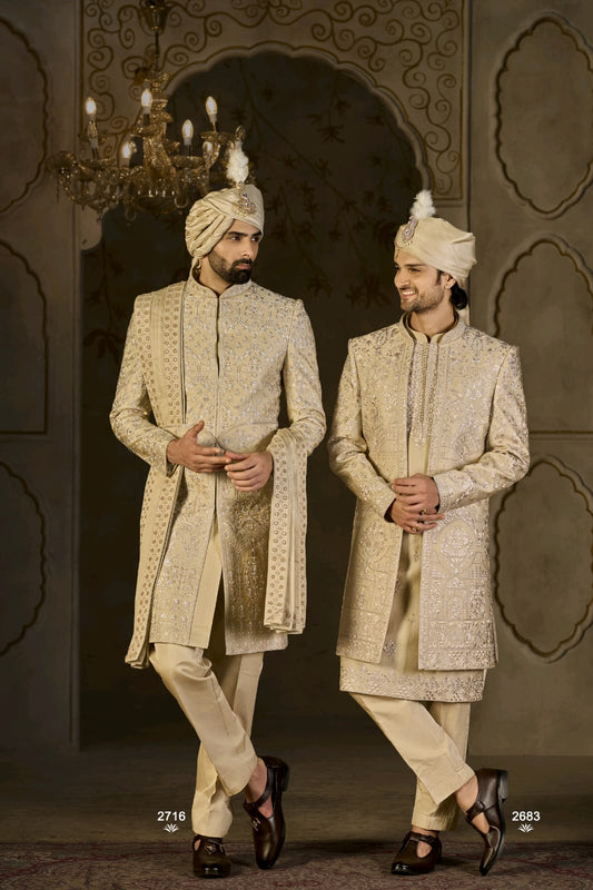 Two men in traditional embroidered sherwanis standing against an ornate wall.