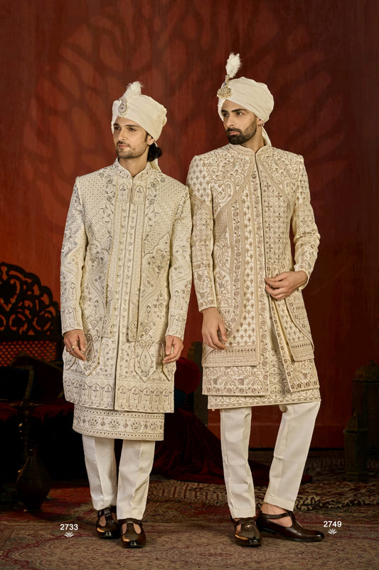 Two men in traditional embroidered sherwanis standing against a patterned red background.