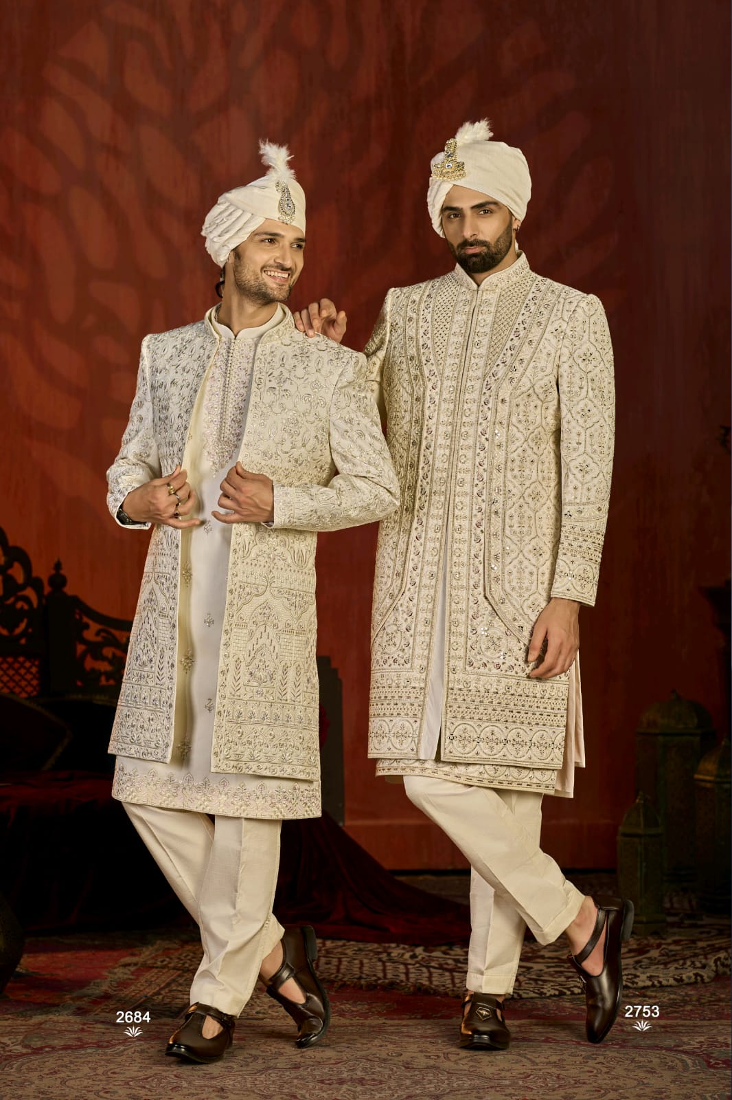 Two men in traditional embroidered sherwanis standing against a red background